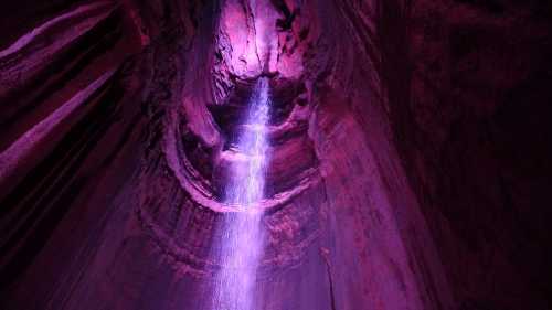 A vibrant purple-lit cave with a waterfall cascading down from the ceiling, surrounded by textured rock formations.