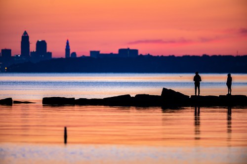 Silhouettes of two people on rocks by a calm lake at sunset, with a city skyline in the background.