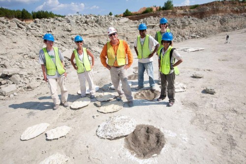 A group of six workers in hard hats and vests stands in a quarry, posing near large circular impressions in the ground.