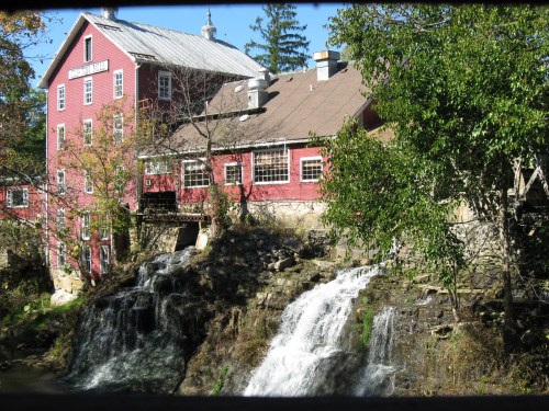A red mill building beside a rocky waterfall, surrounded by trees and greenery in a serene outdoor setting.
