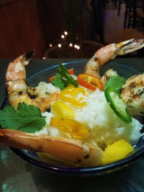 A bowl of grilled shrimp on a bed of rice, garnished with mango, cilantro, and sliced peppers.