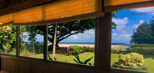 View from a window showcasing a beach, lush greenery, and a clear blue sky with scattered clouds.