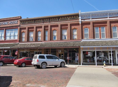 Historic brick buildings line a street, featuring shops and parked cars under a clear blue sky.