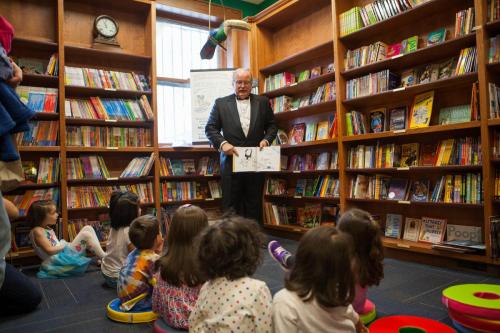 An elderly man in a tuxedo reads to a group of children seated on the floor in a colorful library. Bookshelves are filled with books.