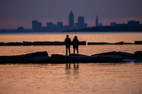 Silhouetted couple standing on rocks by the water, with a city skyline at sunset in the background.