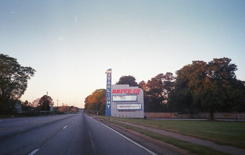 A vintage drive-in theater sign at sunset, with trees lining the roadside and a clear sky above.