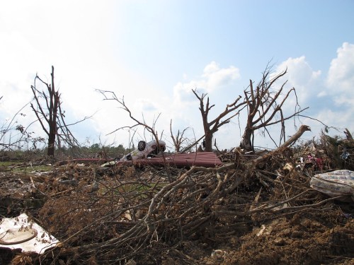 A devastated landscape with uprooted trees, debris, and a flipped vehicle under a cloudy sky.