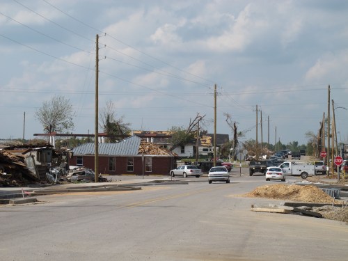 A street scene showing damaged buildings and debris after a storm, with cars and utility poles lining the road.