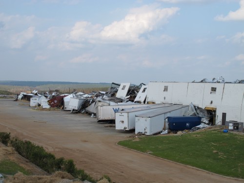 A damaged warehouse with overturned trailers and debris scattered across a dirt lot under a cloudy sky.