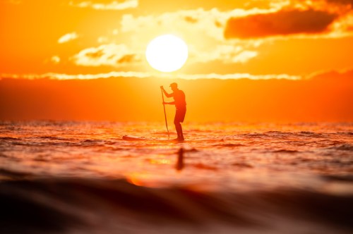 A silhouette of a person paddleboarding at sunset, with a vibrant orange sun setting over the ocean.