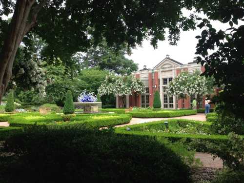 A lush garden with manicured hedges, flowering trees, and a fountain in front of a classic building.