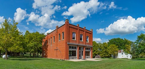 A red brick building with large windows, surrounded by green grass and trees, under a blue sky with fluffy clouds.