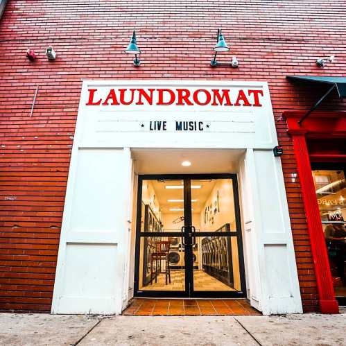 Entrance of a laundromat with a sign for live music, featuring a red brick exterior and glass doors.