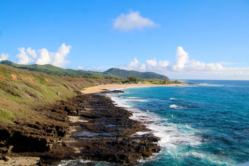 Scenic coastal view with rocky shoreline, sandy beach, and lush green hills under a blue sky with fluffy clouds.