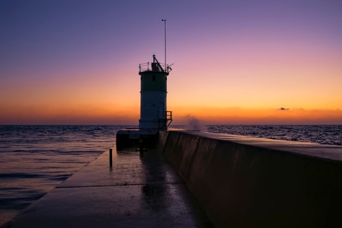 Silhouette of a lighthouse on a pier at sunset, with waves splashing against the structure and a colorful sky.