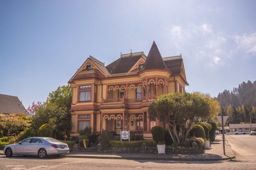 Victorian-style house with intricate details, surrounded by greenery and a parked car in a sunny setting.