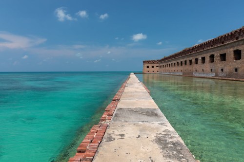 A stone pathway leads to a historic brick fort surrounded by clear turquoise waters under a blue sky.