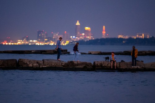 People walking on a rocky pier at dusk, with a city skyline illuminated in the background.