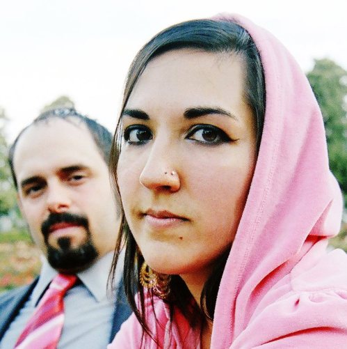 A woman in a pink hoodie gazes at the camera, while a man in a suit is blurred in the background.