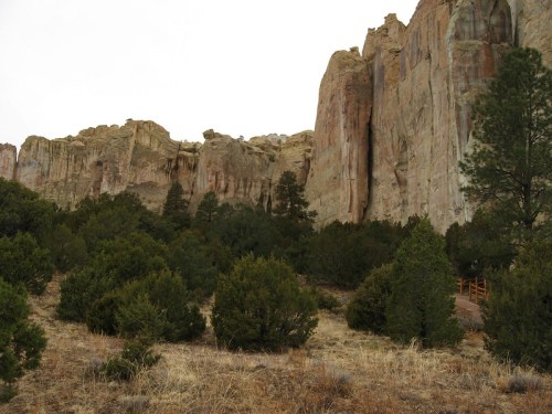 Rocky cliffs rise above a dense area of green shrubs and trees, set against a cloudy sky.