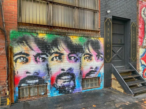 Colorful street art featuring three overlapping faces on a brick wall beside a tattoo shop entrance.