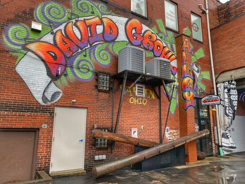 Colorful graffiti reading "David Grohl" on a brick wall, with air conditioning units and wooden logs in front.