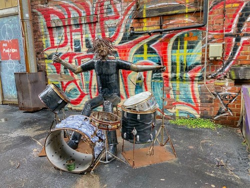 A metal sculpture of a drummer playing on a set of drums, set against a colorful graffiti wall.