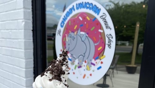 A close-up of a soft serve ice cream cone with crushed cookies, in front of a colorful donut shop sign.