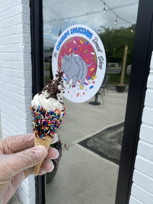 A hand holding an ice cream cone topped with sprinkles and crushed cookies, in front of a colorful donut shop window.