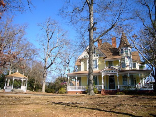 A large Victorian-style house with a gazebo in a bare yard, surrounded by trees under a clear blue sky.
