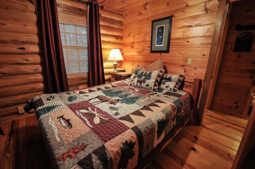 Cozy log cabin bedroom with a patterned quilt, wooden walls, a lamp, and a window with natural light.
