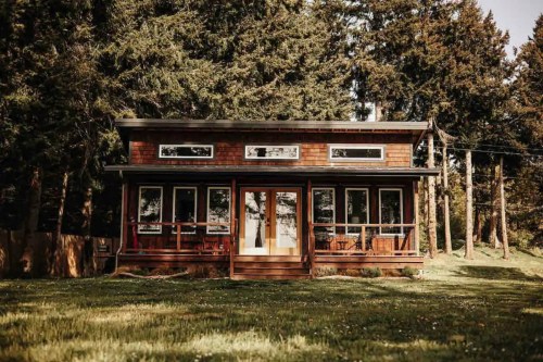 A cozy wooden cabin surrounded by tall trees, featuring large windows and a welcoming porch.