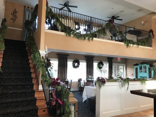 A festive interior with garlands and wreaths, featuring a staircase leading to a dining area with tables set for a celebration.