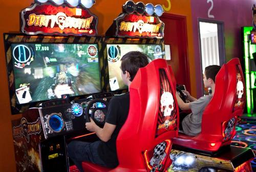 Two kids playing racing arcade games in a colorful game room, focused on their screens.