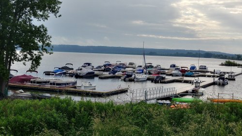 A marina with various boats docked on a calm lake, surrounded by greenery and cloudy skies.