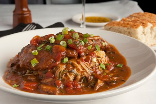 A plate of chicken topped with green onions, served over noodles in a rich sauce, with bread in the background.