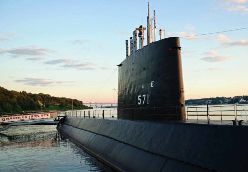 A submarine docked at sunset, with its bow facing the camera and a calm water surface reflecting the sky.