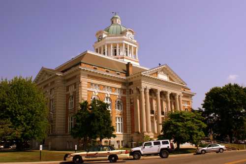 Historic courthouse with a green dome, tall columns, and surrounding trees, set against a clear blue sky.