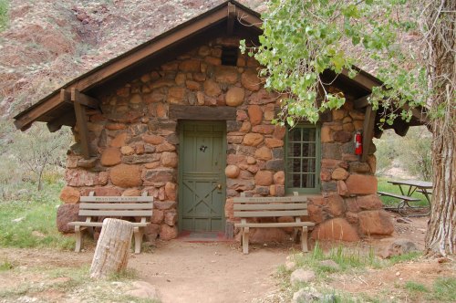 A rustic stone cabin with a green door, surrounded by trees and benches, set in a natural landscape.