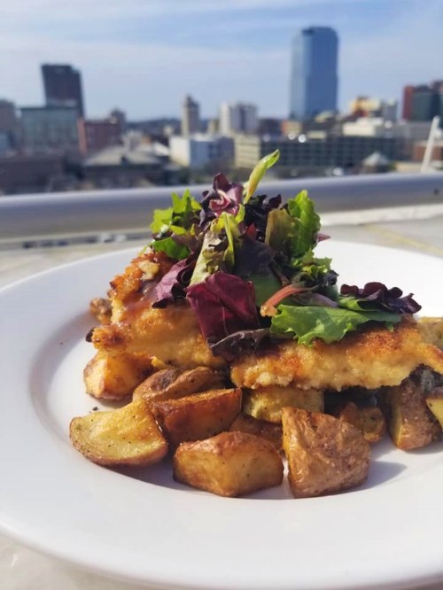 A plate of crispy fish topped with fresh greens, served with roasted potatoes, against a city skyline backdrop.