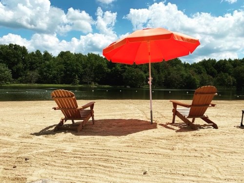 Two wooden chairs under an orange umbrella on a sandy beach by a calm lake, surrounded by green trees and blue skies.