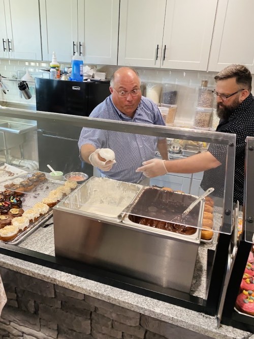 A man in a blue shirt serves ice cream while another man in a black shirt assists in a bakery setting.