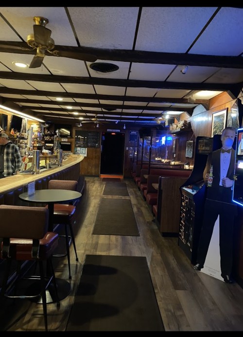 A dimly lit bar interior with a long counter, seating booths, and a cardboard cutout of a tuxedoed man.