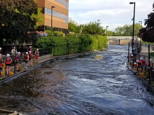 A group of people in life jackets working along a flowing river near a building, with trees and greenery in the background.