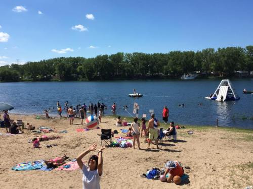 A sunny beach scene with people playing, swimming, and relaxing by a lake, featuring a water slide and beach balls.