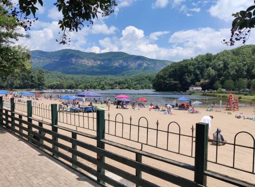 A sandy beach with umbrellas and people enjoying the sun, surrounded by mountains and a calm lake under a blue sky.