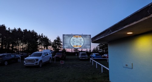 A drive-in movie screen displays "Tune to Film Union 100.5 FM" at dusk, with parked cars in the foreground.