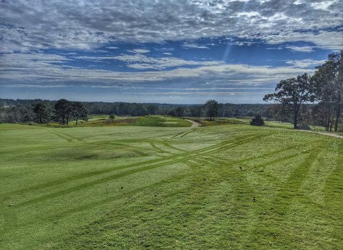A scenic view of a golf course with green grass, tire tracks, and a cloudy blue sky in the background.