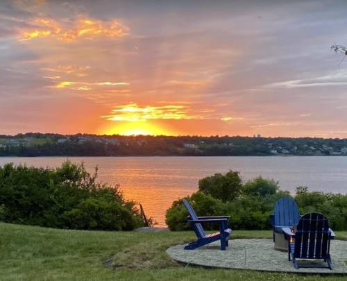 A serene sunset over a lake, with two blue chairs facing the water and lush greenery in the foreground.
