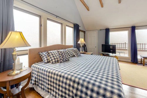 Cozy bedroom with a large bed, blue checkered bedding, lamps, and a TV, featuring large windows and wooden beams.
